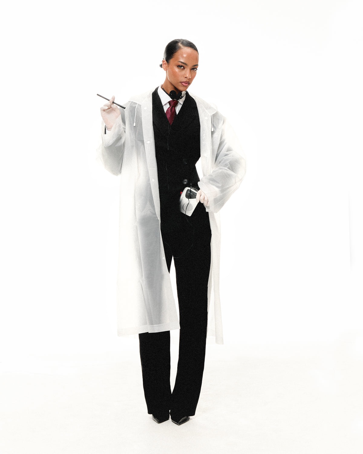 Model in a clear lab coat over a black suit, holding a lip pencil